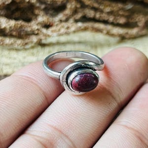 May include: A silver ring featuring a dark red oval gemstone. The ring has a distinctive, swirling pattern around the stone. The ring is held by a person's fingers.