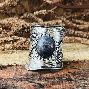 Peut inclure: Bague argentée ornée d'une grosse pierre ovale bleu-gris au centre. La bague présente un motif floral texturé autour de la pierre et sur le corps. La bague est présentée sur une surface en bois.
