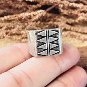 May include: A silver-toned ring with a rectangular face. The face is decorated with a repeating pattern of triangles. The ring is held by a hand.