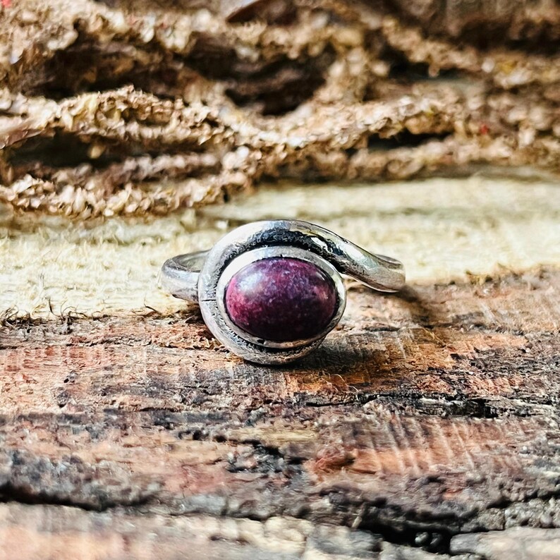 May include: A silver ring featuring a large, oval, deep red gemstone. The ring's band has a swirling design that wraps around the gemstone. The ring is set against a rustic wooden background.