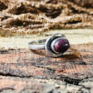 May include: A silver ring featuring a dark red, oval gemstone. The stone is set within a circular design with a textured outer band. The ring rests on a textured, brown surface.