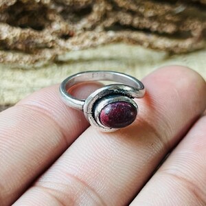May include: A silver ring featuring a dark red oval gemstone. The ring has a distinctive, swirling pattern around the stone. The ring is held by a person's fingers.