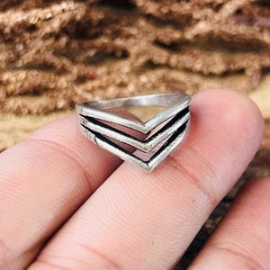 May include: A silver-toned ring with a chevron design. The ring features three stacked V-shaped layers, with dark lines accentuating the edges. The ring is held on a person's finger.