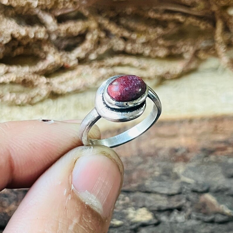 May include: A silver ring featuring a large, oval, reddish-purple stone. The stone is set in a swirl design. The ring is held by a person's fingers, with a wooden background.