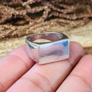 May include: A silver ring with a rectangular face. The ring is worn on a finger. The ring has a slightly worn appearance, suggesting it may be vintage or antique. The ring is a simple, classic design.