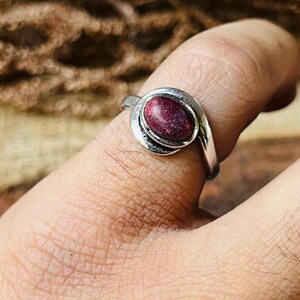 May include: A silver ring with a swirling design, featuring a large, oval-shaped, deep red gemstone. The ring is shown on a finger, with the focus on the jewelry's details and craftsmanship. The gemstone has a textured appearance.