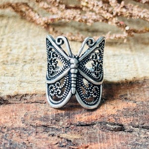 May include: A silver-colored butterfly ring with intricate filigree detailing. The ring features a detailed butterfly design with openwork wings and a textured body. The ring is set against a rustic wooden surface.