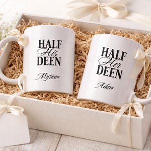 Könnte beinhalten: Zwei weiße Tassen mit den Aufschriften "HALF His DEEN" und "HALF Her DEEN" in schwarzer Schrift, mit Namen darunter, befinden sich in einer Geschenkbox. Die Box ist mit Holzwolle gefüllt und mit Schleifen verziert.