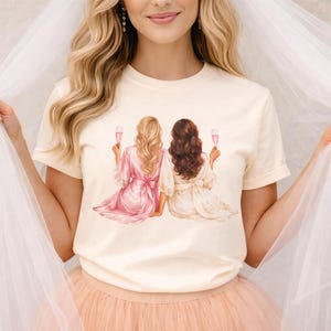 May include: A cream-coloured t-shirt with an illustration of two figures in robes, holding champagne flutes. One figure has blonde hair and a pink robe, the other has brown hair and a cream-coloured robe. The figures are seated, with their backs to the viewer.