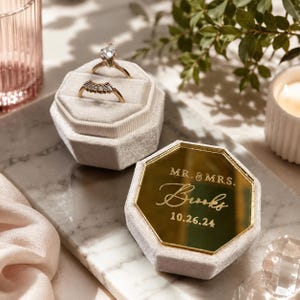 May include: A gold engagement ring with a large diamond and a gold wedding band rest in a light gray velvet ring box. A second ring box with a gold mirrored top displays the text "MR. & MRS. Brooks 10.26.24".