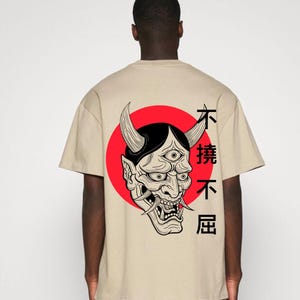 Puede incluir: Camiseta beige con un diseño de máscara Oni en blanco y negro sobre un círculo rojo. La máscara tiene cuernos y múltiples ojos. Caracteres japoneses impresos a la derecha. El texto "Leravia Studio Art" está impreso en la parte superior.
