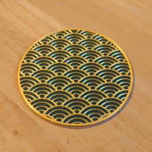 Puede incluir: Un posavasos redondo con un borde dorado y un patrón de olas repetidas en verde azulado oscuro. El posavasos está hecho de un material resistente y está diseñado para proteger las superficies de la condensación de las bebidas.