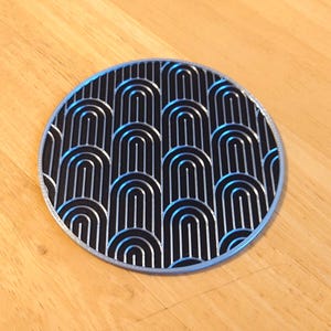 Puede incluir: Un posavasos redondo con fondo negro y diseño art déco plateado. El diseño presenta formas arqueadas repetidas. El posavasos tiene un borde plateado y está sobre una superficie de madera.