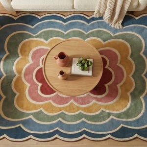 May include: Overhead shot of a multi-coloured, scalloped-edge rug with a floral pattern. The rug has layers of blue, green, yellow, pink, and red. A round wooden coffee table with decor is in the centre. Two woven chairs are on the left.