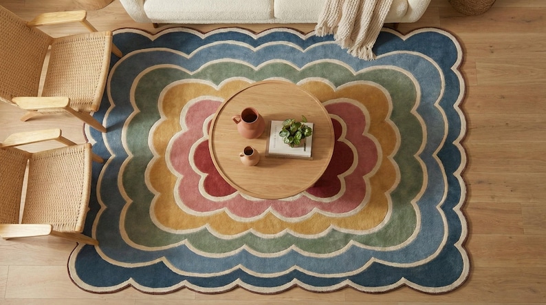 May include: Overhead shot of a multi-coloured, scalloped-edge rug with a floral pattern. The rug has layers of blue, green, yellow, pink, and red. A round wooden coffee table with decor is in the centre. Two woven chairs are on the left.