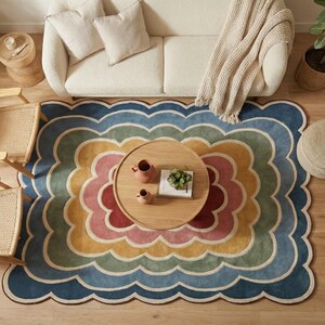 May include: An overhead shot of a living room featuring a multi-coloured, scalloped-edge rug. The rug has a rainbow design with blue, yellow, and pink hues. A wooden coffee table, two chairs, and a sofa are arranged on the wooden floor.