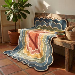 May include: A multi-coloured, scalloped-edge rug in shades of blue, green, yellow, pink, and orange. The rug is draped over a wooden bench, with two ceramic bowls and a basket of yarn nearby. A potted plant is in the background.
