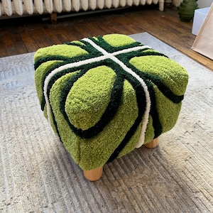 May include: Green and white patterned ottoman with wooden legs. The ottoman is made of a soft, plush fabric.