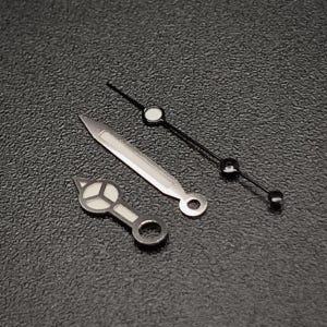 Puede incluir: Primer plano de agujas y componentes de reloj. La imagen muestra tres agujas de reloj: una aguja horaria plateada, una aguja minutera plateada y un segundero negro con detalles blancos y negros. Las agujas están sobre una superficie negra texturizada.