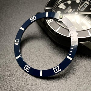 Pode incluir: Uma luneta de relógio azul marinho com algarismos e marcadores brancos, exibindo os números de 0 a 50. A luneta é circular e fica em frente a um mostrador de relógio preto com o logotipo Seiko.