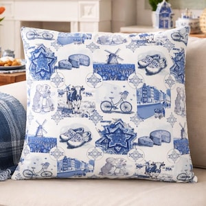 Op de afbeelding: Een vierkant decoratief kussen met een Nederlands thema in blauw en wit. Het patroon bevat afbeeldingen van molens, kaas, fietsen, klompen en koeien. Het kussen ligt op een beige bank.