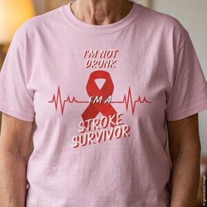 Op de afbeelding: Een lichtroze t-shirt met een rood bewustzijnslintje gecentreerd over een rode hartslaglijn. De in het rood gedrukte tekst luidt I'M NOT DRUNK I'M A STROKE SURVIVOR. Het ontwerp bevindt zich op het borstgedeelte van het kledingstuk met korte mouwen.