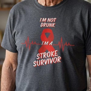 Pode incluir: Uma t-shirt cinzenta escura de manga curta com um laço vermelho de consciencialização centrado sobre um gráfico de linha de batimento cardíaco vermelho. O texto I'M NOT DRUNK I'M A STROKE SURVIVOR está em letras brancas acima e abaixo.