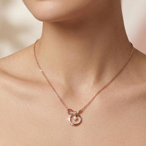 Peut inclure: Un collier en or rose avec un nœud délicat et un pendentif circulaire orné de petits diamants étincelants. Le collier est porté par une personne vêtue d'un haut rose pâle.