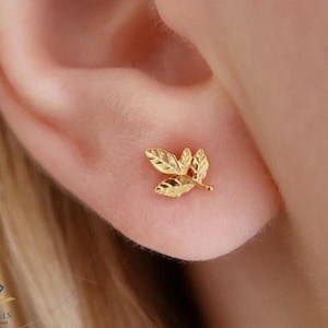 Peut inclure: Une boucle d'oreille en forme de feuille dorée est présentée sur une oreille. La boucle d'oreille a un design détaillé de trois feuilles. La boucle d'oreille est petite et délicate, et la couleur dorée lui donne un aspect chaleureux et élégant. C'est un bijou.