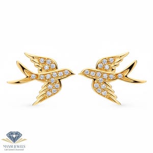 Puede incluir: Un par de pendientes dorados con forma de golondrinas, cada uno adornado con pequeñas y brillantes gemas blancas. Los pendientes están sobre un fondo blanco, mostrando el diseño y la artesanía de la joyería.