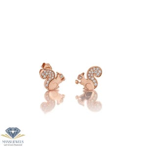 May include: A pair of rose gold squirrel stud earrings. Each earring features a squirrel design with a textured body and a fluffy tail, embellished with small, sparkling stones. The earrings are set against a white background.