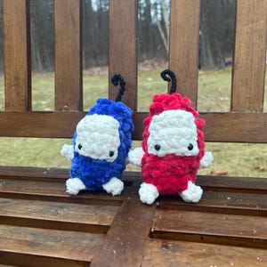 May include: Two handmade crochet figures, one blue and one red, each with a white face and small arms and legs. They have black curly stems on top. The figures are sitting on a wooden bench.