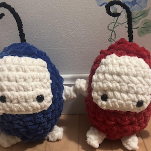 May include: Two plush toys, one blue and one red, are displayed. Each has a white face, small black eyes, and a black antenna. The toys are made of a soft, textured material, and are likely handmade. They are positioned on a wooden surface.
