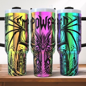 May include: Three stainless steel tumblers with a gradient rainbow finish. Each tumbler features a black dragon and castle design with the words "EMPOWERED" in a gothic font. The tumblers have a white lid and a black handle.