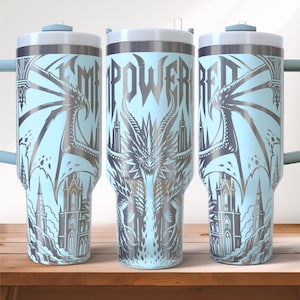 May include: A light blue tumbler with a silver dragon and castle design. The word "EMPOWERED" is displayed vertically. The tumbler has a silver rim, a white lid, and a handle.