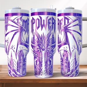 May include: White tumbler with a purple dragon design and the word "EMPOWERED" in purple. The design includes a dragon, castle, and architectural elements. The tumbler has a white lid and a purple rim.