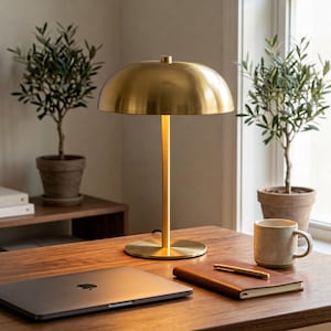 May include: A brushed brass table lamp with a dome-shaped shade and a slender cylindrical stem on a circular base. The lamp features a metallic gold finish and is positioned on a wooden surface alongside a laptop, a notebook, and a ceramic mug.