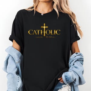 May include: Black t-shirt featuring the word "CATHOLIC" in gold, with a gold cross above it. Below the word is "est. 33 AD". The shirt is a casual style. The text and cross are in a gold serif font.