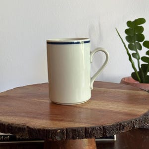 May include: A cylindrical off-white ceramic mug featuring a single thin navy blue horizontal stripe encircling the upper rim. The mug has a smooth finish, a straight body, and a curved handle extending from the side for a comfortable grip.