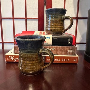 May include: Two handmade ceramic mugs with a blue rim and handle, and a brown and tan body. The mugs are sitting on top of a stack of books with red and brown covers. The mugs have a rustic, textured appearance.