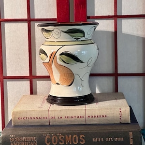 Könnte beinhalten: Eine dekorative Keramikvase mit weißem Sockel, verziert mit grünen und braunen Blatt- und Fruchtdesigns. Die Vase steht auf einem Stapel von Büchern, darunter "DICTIONNAIRE DE LA PEINTURE MODERNE" und "the COSMOS".