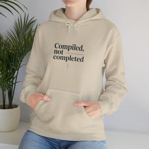 May include: A beige hoodie with a front pocket and a drawstring hood. The text "Compiled, not completed" is printed in black on the front. The person is wearing blue jeans.