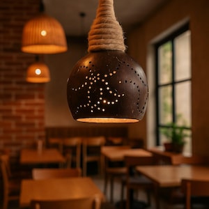 May include: A dark brown pendant light with a spherical shape and numerous small holes, emitting a warm glow. The light is suspended by a thick, natural rope. Other lights are visible in the background, along with tables and chairs, suggesting a restaurant setting.