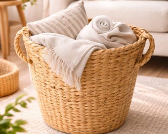 May include: A large, round woven basket with two handles, filled with folded blankets and a striped pillow. The basket is light brown and sits on a neutral-colored rug, suggesting a cozy home decor theme.