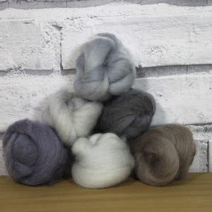 Op de afbeelding: Een verzameling wolvlies in verschillende neutrale kleuren, waaronder grijs, wit en bruin, gerangschikt in een piramidevorm. De zachte, donzige wol is perfect voor handwerkprojecten. De achtergrond is een witte bakstenen muur.