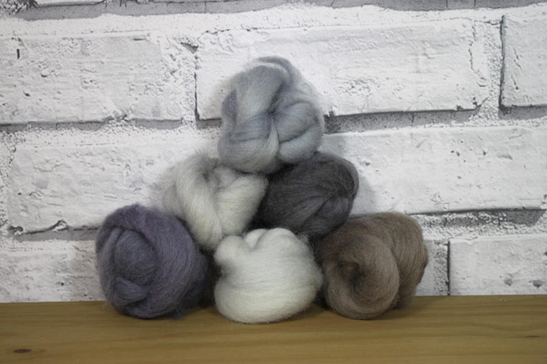 Op de afbeelding: Een verzameling wolvlies in verschillende neutrale kleuren, waaronder grijs, wit en bruin, gerangschikt in een piramidevorm. De zachte, donzige wol is perfect voor handwerkprojecten. De achtergrond is een witte bakstenen muur.