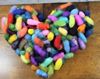 Könnte beinhalten: Eine herzförmige Anordnung von bunten gefilzten Wollrohlingen. Die Rohlinge sind in verschiedenen Schattierungen von Rot, Orange, Gelb, Grün, Blau, Lila und Rosa.