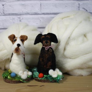 Könnte beinhalten: Zwei Nadelfilz-Hundefiguren, eine weiß und braun mit einem rot-weißen Halsband, die andere schwarz und braun mit einem rosa-weißen Halsband, sitzen auf einem grünen Sockel mit einem roten und blauen Ball. Die Figuren stehen vor zwei Knäueln weißer Wolle.