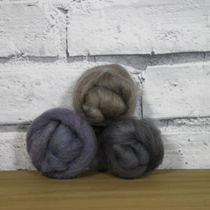 Op de afbeelding: Drie bollen wol in grijstinten, gerangschikt in een driehoekige compositie. De wolbollen zijn diepgrijs, lichtgrijs en taupe. De achtergrond is een witte bakstenen muur en een houten oppervlak.