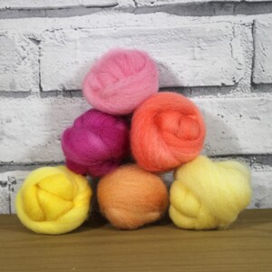May include: Six balls of wool yarn in various shades of pink, orange, and yellow. The balls are arranged in a pyramid shape.